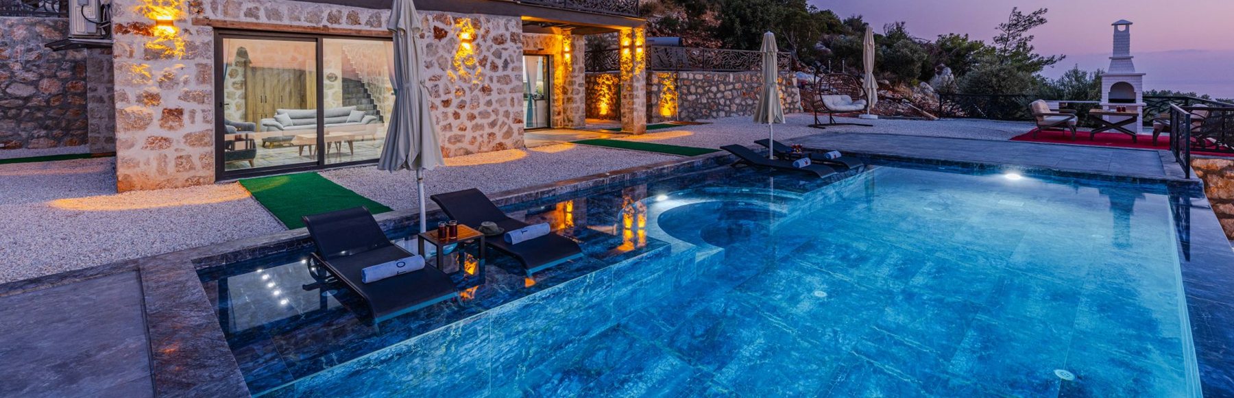 Swimming pool and shallow loungers beside a stone building at dusk.