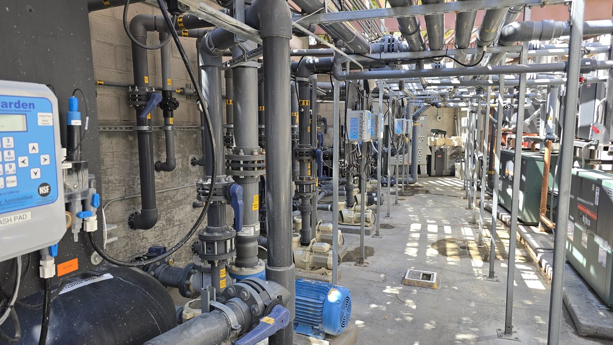 Commercial pool equipment area with chemical controllers mounted beside rows of piping and pumps.